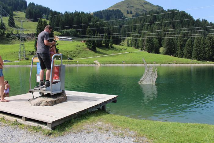 En man och ett barn står på en liten träbrygga vid en sjö i Westendorf och använder en myntdriven maskin. I sjön dyker en drakeskulptur upp ur vattnet, omgiven av gröna kullar och tallar – perfekt för sommarferie Österrig.