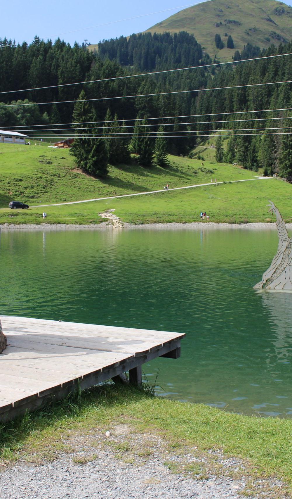 En man och ett barn står på en liten träbrygga vid en sjö i Westendorf och använder en myntdriven maskin. I sjön dyker en drakeskulptur upp ur vattnet, omgiven av gröna kullar och tallar – perfekt för sommarferie Österrig.