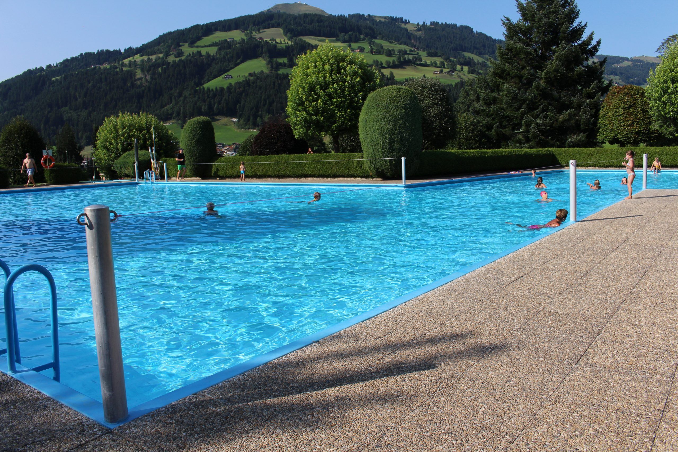 En lys udendørs swimmingpool med folk, der nyder vandet på en solskinsdag under deres sommerferie i Westendorf. Poolen er omgivet af en pænt trimmet hæk og træer, med bølgende grønne bakker og et bjerg under en klar blå himmel i baggrunden.