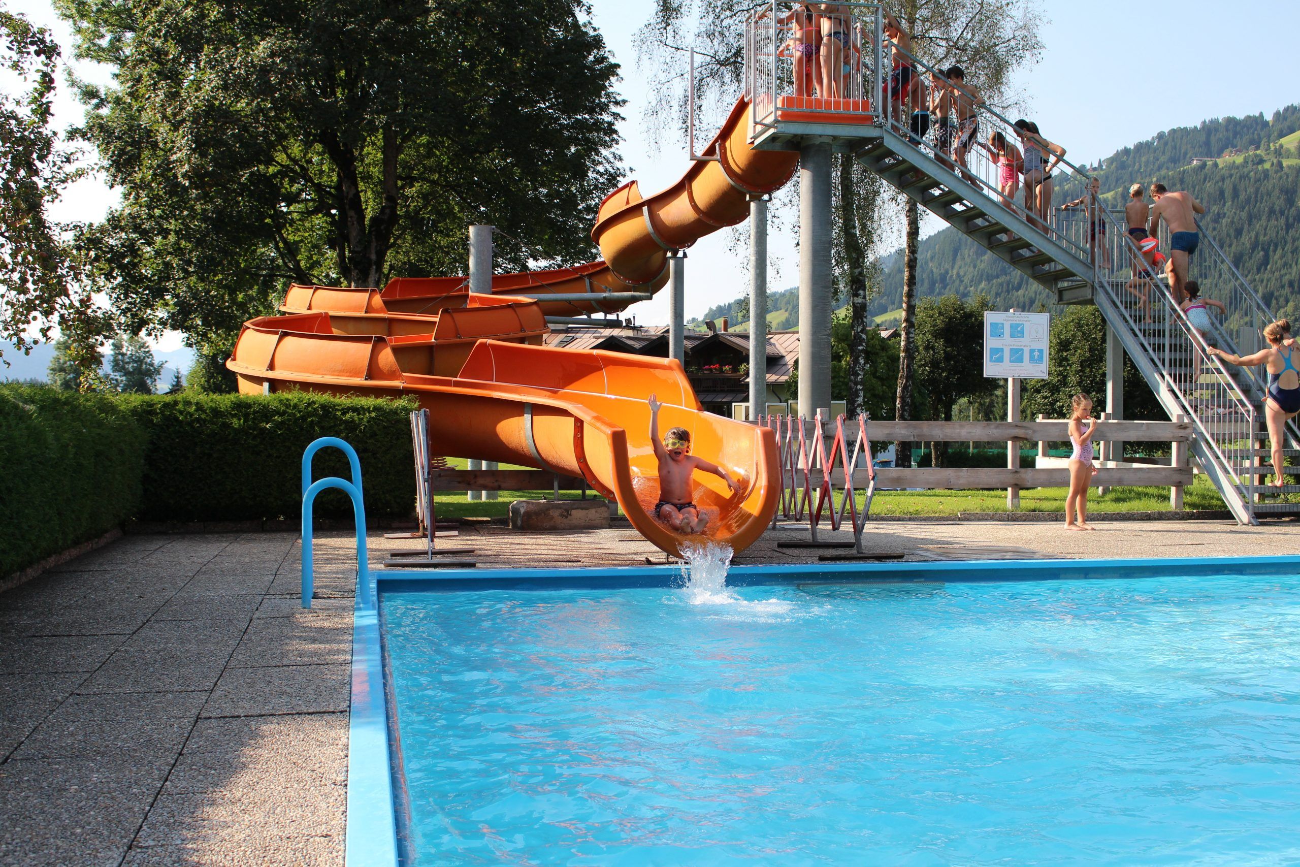 Midt i en solskinsdag perfekt til sommerferie i Westendorf glider en person yndefuldt ned ad den orange vandrutsjebane ned i den indbydende klarblå pool. Ivrige børn venter på deres tur på toppen, mens den naturskønne kulisse af træer og bakker fuldender den pulserende scene.