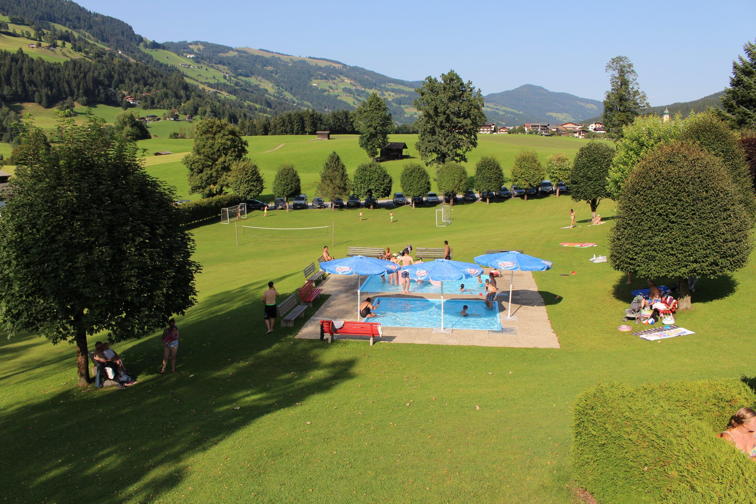 Människor kopplar av och leker runt en liten utomhuspool med blå parasoller på ett stort gräsbevuxet fält i Westendorf, omgivet av träd och berg under en klar himmel – perfekt för en sommarferie i Österrike eller minnesvärda skidresor under alla årstider.