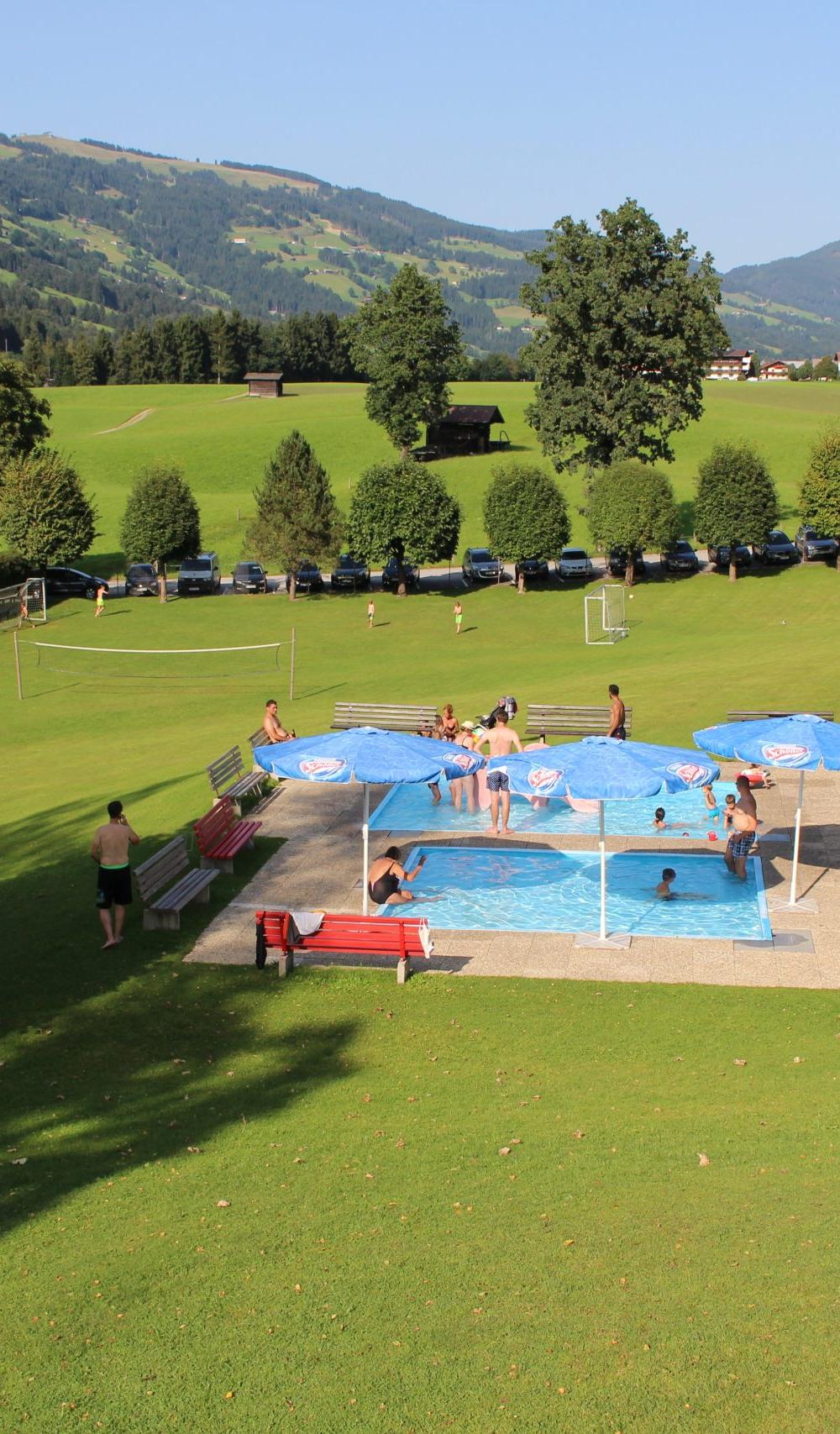 Människor kopplar av och leker runt en liten utomhuspool med blå parasoller på ett stort gräsbevuxet fält i Westendorf, omgivet av träd och berg under en klar himmel – perfekt för en sommarferie i Österrike eller minnesvärda skidresor under alla årstider.