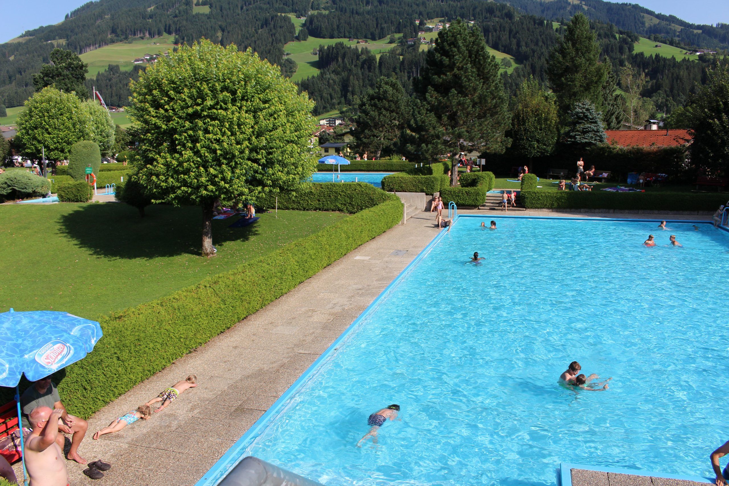 Under en solrig sommerferie i Westendorf er en udendørs swimmingpool omgivet af grønne hække og træer, hvor folk svømmer og soler sig. I baggrunden rejser sig frodige grønne bakker bag rekreative paraplyer.