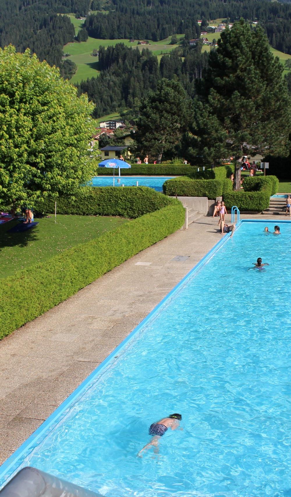 Under en solrig sommerferie i Westendorf er en udendørs swimmingpool omgivet af grønne hække og træer, hvor folk svømmer og soler sig. I baggrunden rejser sig frodige grønne bakker bag rekreative paraplyer.