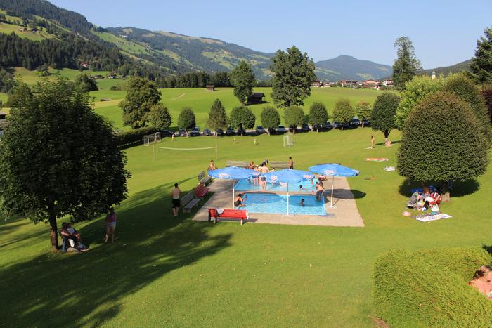 En naturskøn park med en lille pool, blå parasoller og mennesker, der slapper af, fanger essensen af en Sommerferie i Westendorf. Det græsklædte område er oversået med træer mod bølgende bakker og en fodboldbane, mens besøgende slapper af på græsset og nyder den solrige dag.