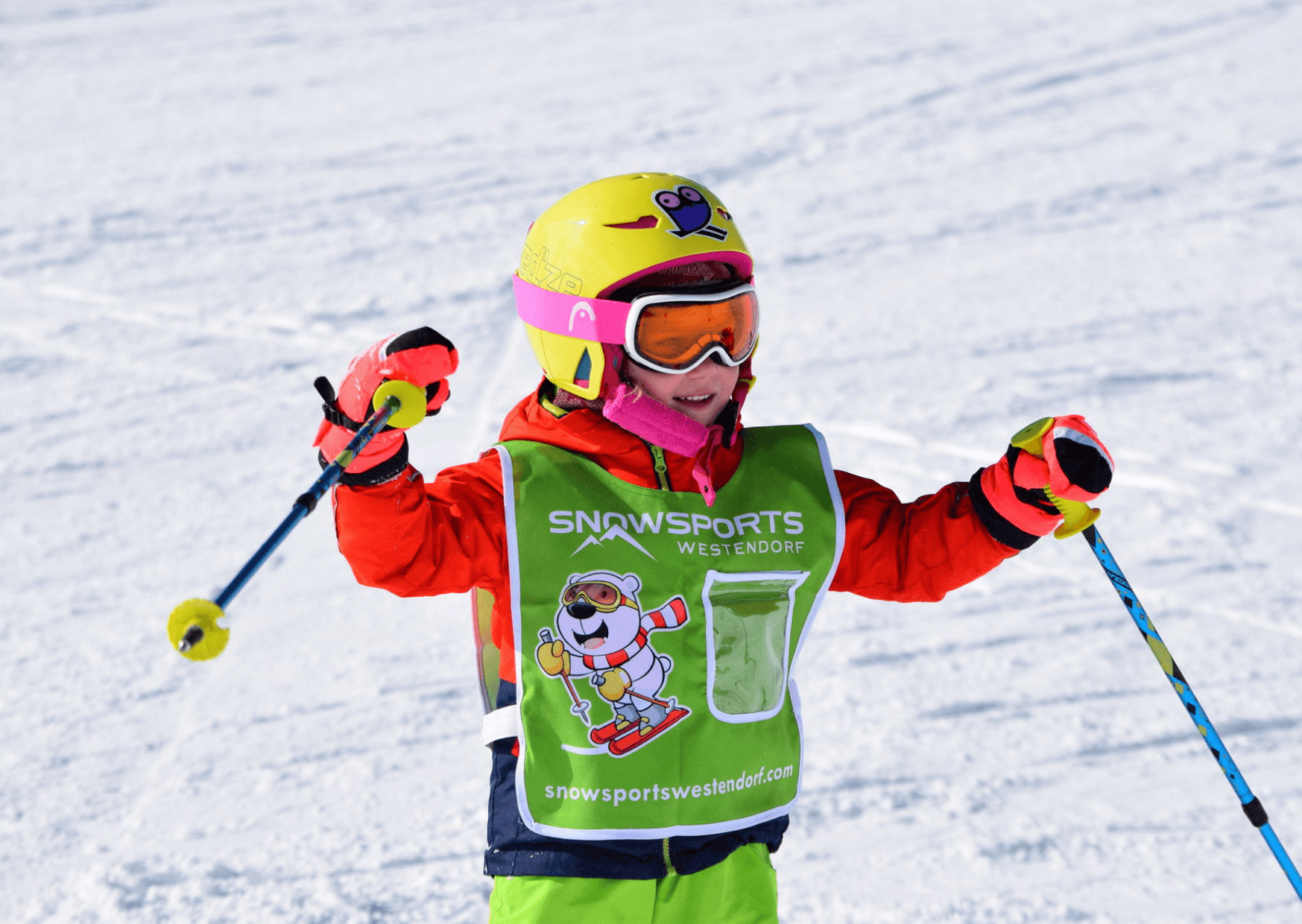 Et barn i farverigt skiudstyr, inklusive en gul hjelm og orange jakke, står på ski på sne. De holder skistave og har en vest på, hvor der står Snowsports Westendorf. Denne livlige scene fanger skirejsers glæde på baggrund af et snedækket landskab.