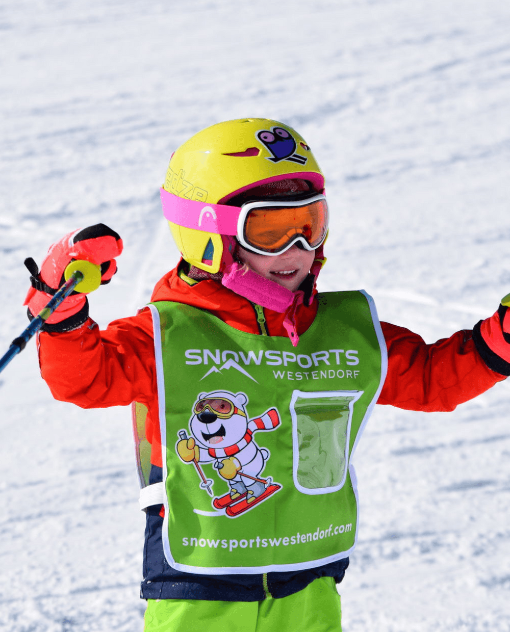 Et barn i farverigt skiudstyr, inklusive en gul hjelm og orange jakke, står på ski på sne. De holder skistave og har en vest på, hvor der står Snowsports Westendorf. Denne livlige scene fanger skirejsers glæde på baggrund af et snedækket landskab.