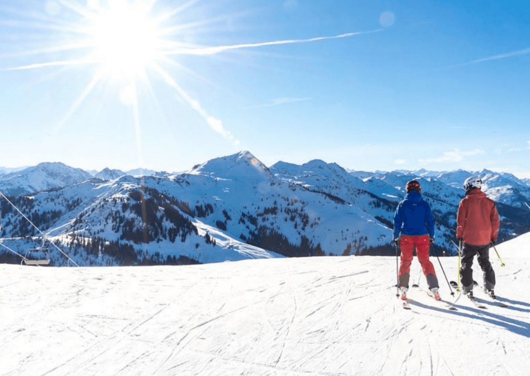 Två skidåkare står på en snöig bergssluttning i Saalbach under en strålande sol, med panoramautsikt över snötäckta toppar och klarblå himmel – en perfekt dag för oförglömliga skidåkningsäventyr.