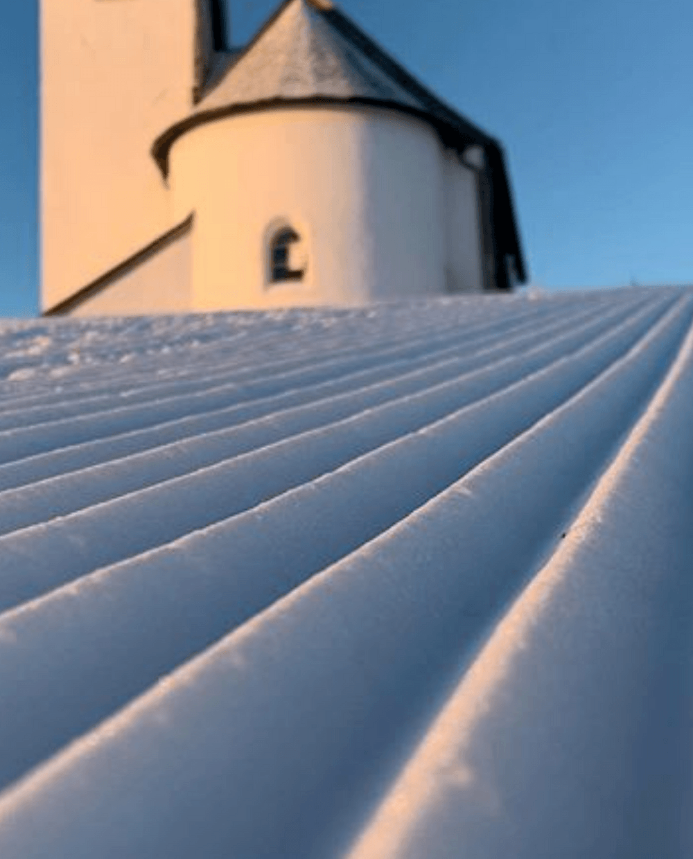 En närbild av nypreparerad snö med parallella linjer som leder mot en solbelyst byggnad med torn och rund sektion mot en klarblå himmel i Saalbach.