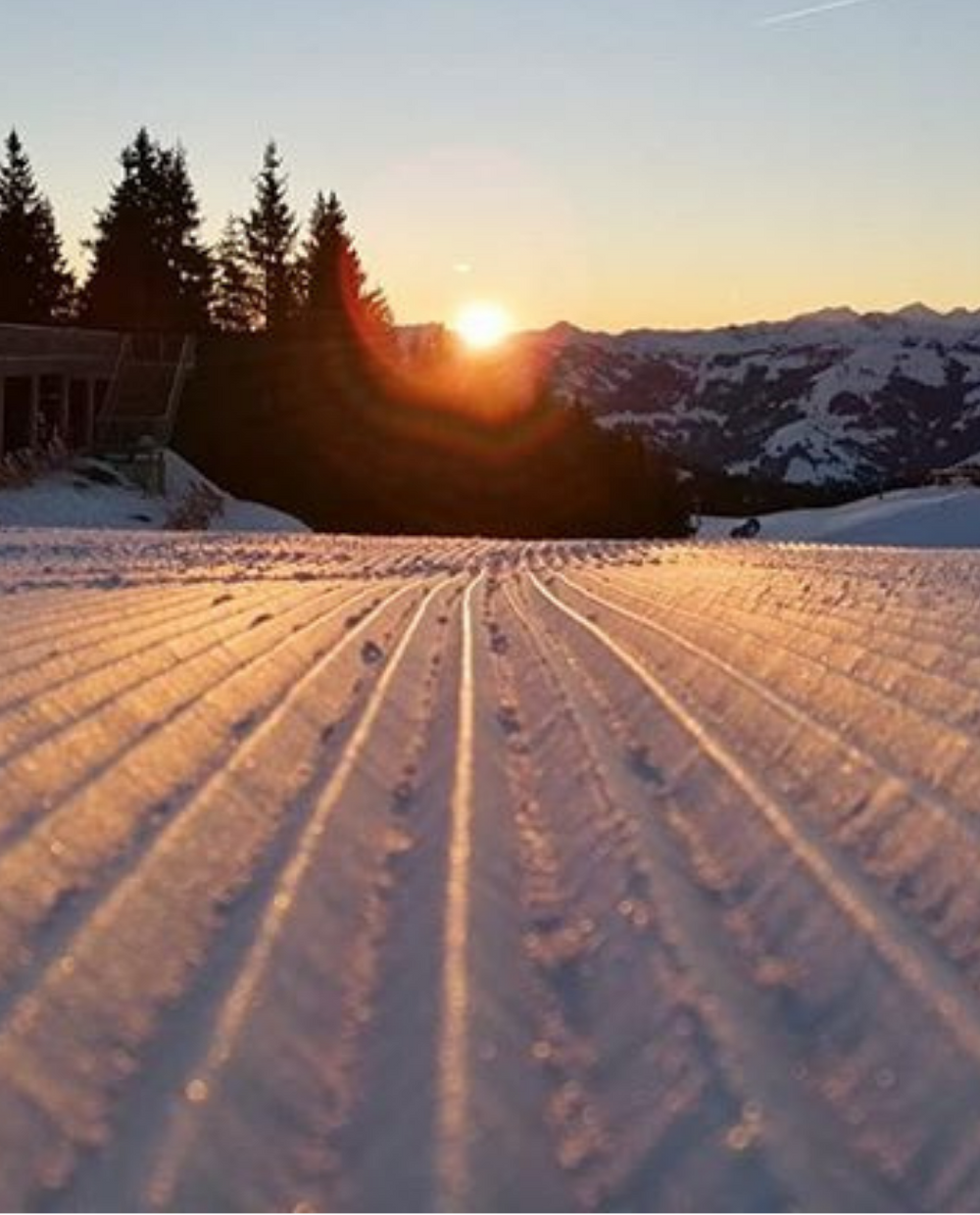 En soluppgång lyser upp nypreparerad snö med parallella linjer, omgivna av tallar och snöklädda berg i bakgrunden. En byggnad ligger till vänster i bilden och frammanar charmen hos en skidåkningsbana i Saalbach.