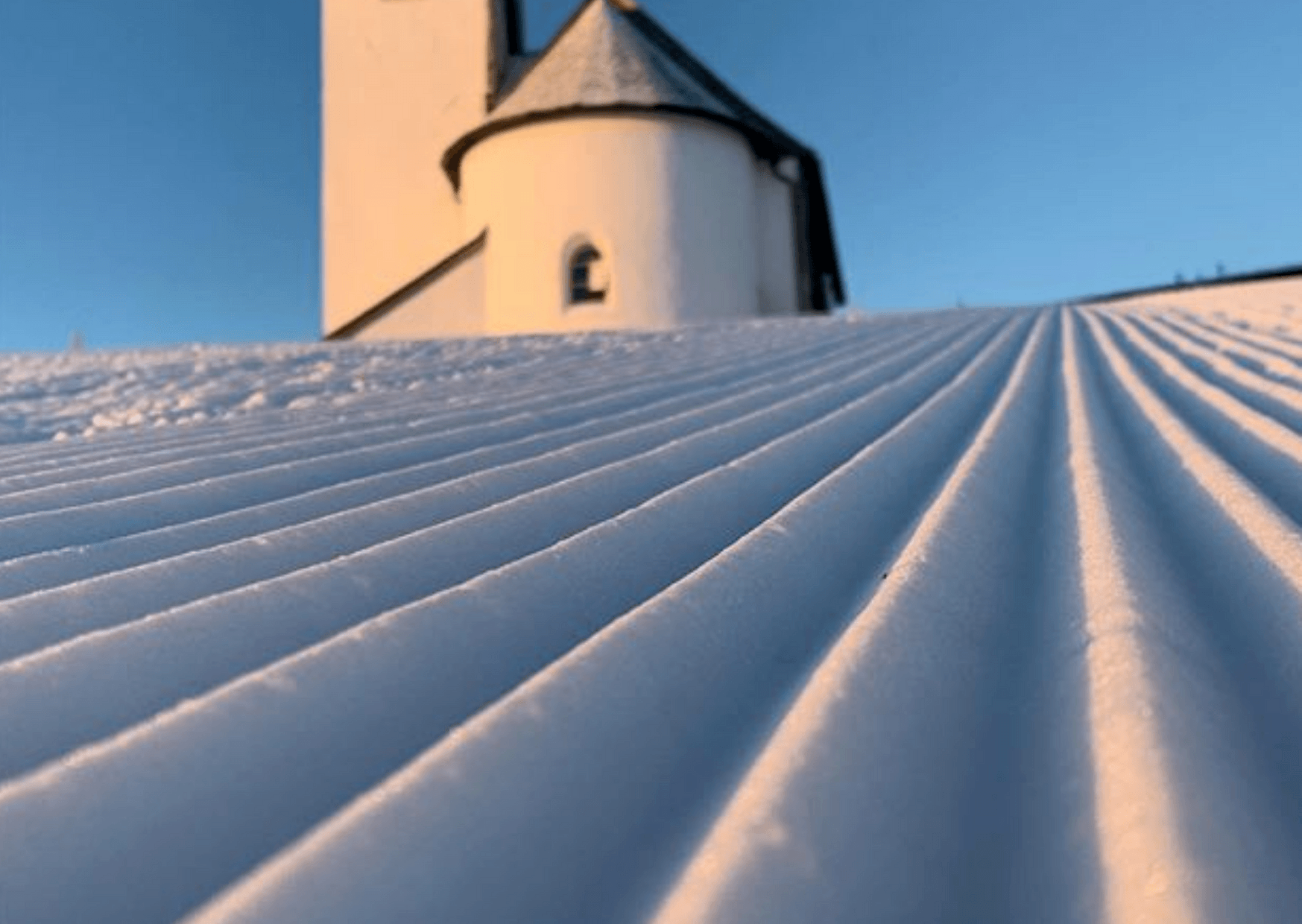 En snedækket skråning med pæne, parallelle riller fører op til en hvid bygning med et lille tårn, sat mod en klar blå himmel - en perfekt scene for din næste skiferie i Westendorf.