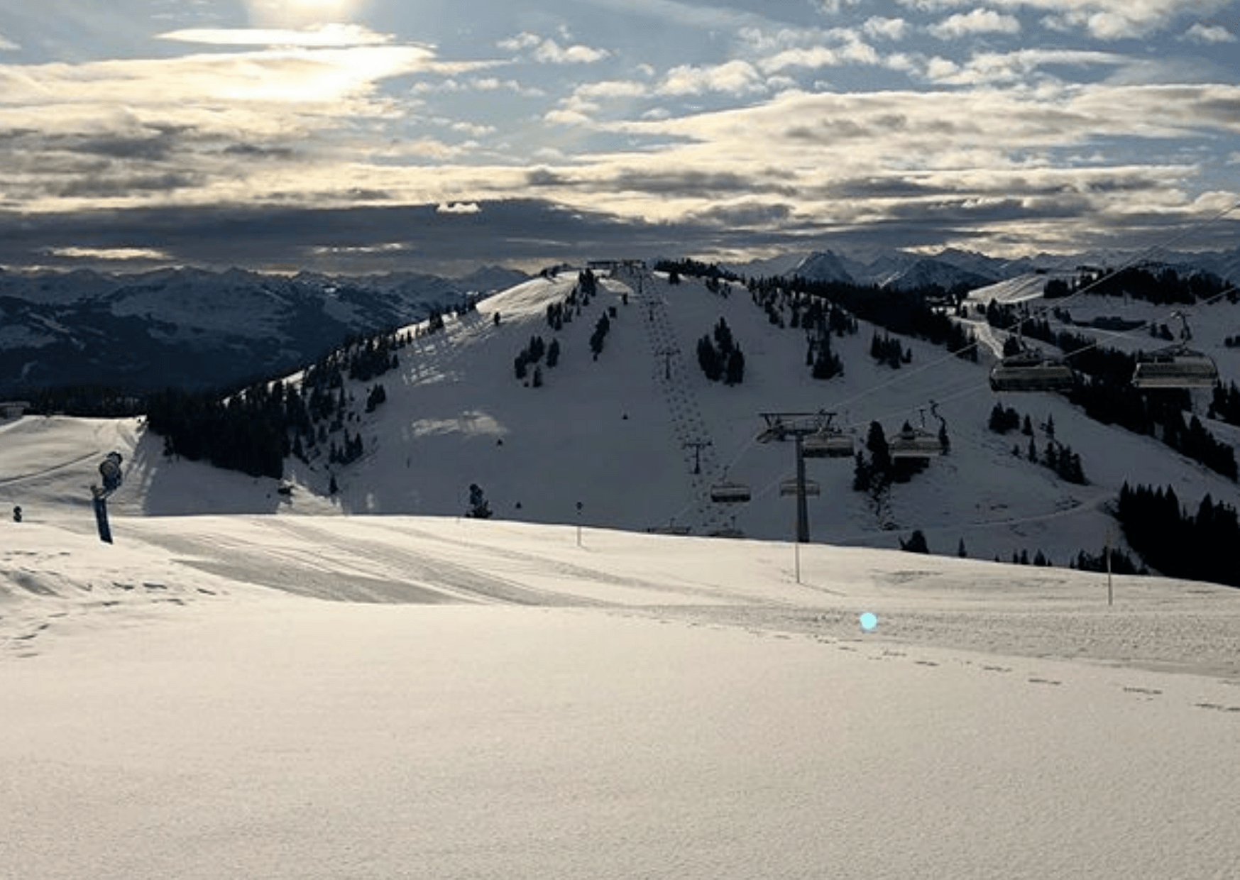 Snedækkede skiløjper i Westendorf under en delvist overskyet himmel, med skilifter langs bakken. Solen kigger gennem skyerne og kaster et blødt lys på det bjergrige landskab i baggrunden – en perfekt ramme om en mindeværdig skiferie.