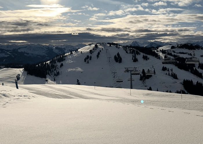 Snedækkede skiløjper i Westendorf under en delvist overskyet himmel, med skilifter langs bakken. Solen kigger gennem skyerne og kaster et blødt lys på det bjergrige landskab i baggrunden – en perfekt ramme om en mindeværdig skiferie.