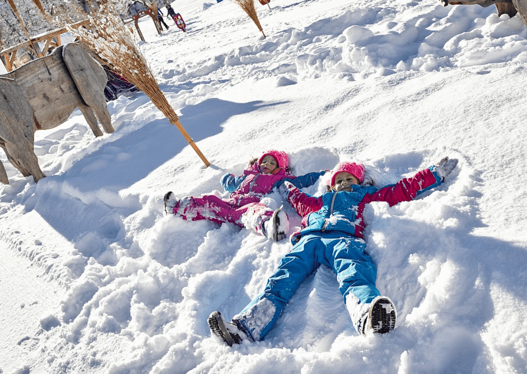 Två barn i färgglada vinterkläder ligger på rygg i snön och gör snöänglar. Marken är täckt av nysnö i Saalbach, med träkonstruktioner och kvastar synliga i bakgrunden.