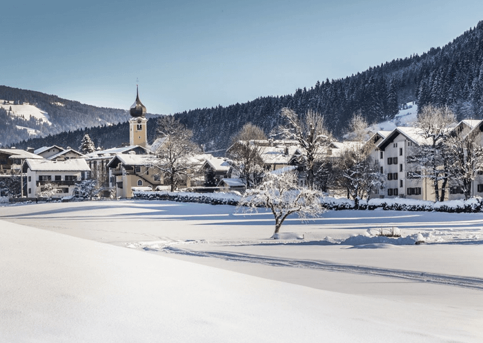 En pittoresk snötäckt by i Saalbach med ett kyrktorn, omgiven av snötäckta hustak, träd och skogsklädda kullar under en klarblå himmel – perfekt för oförglömliga skidresor eller skidåkningsupplevelser.