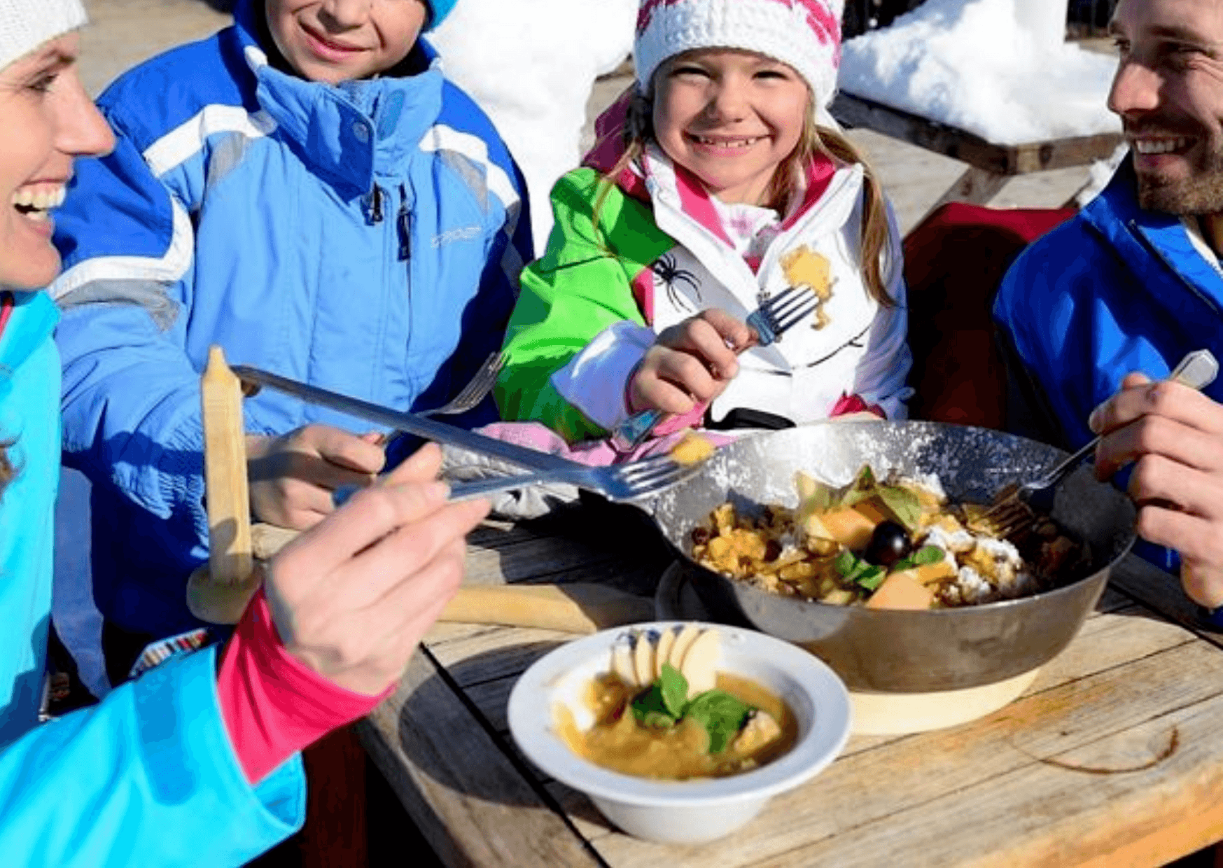 En familie klædt i farverige vinterjakker nyder et måltid udendørs under deres skiferie. De sidder ved et træbord med en pande med kogt mad og en skål suppe. Sne kan ses i baggrunden, og alle smiler og holder redskaber og nyder denne hyggelige stund sammen.