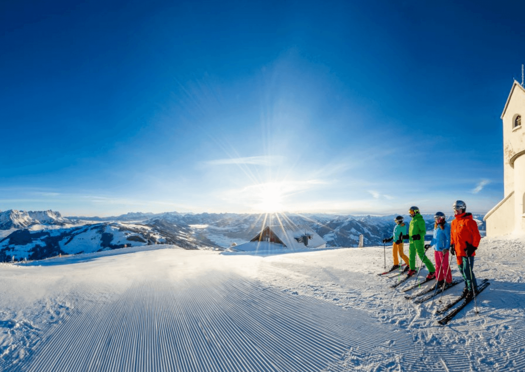 Fyra skidåkare i färgglada kläder står på en preparerad snöig backe med utsikt över bergstoppar och en ljus soluppgång; en liten byggnad syns till höger och fångar magin hos skidresor i Westendorf.
