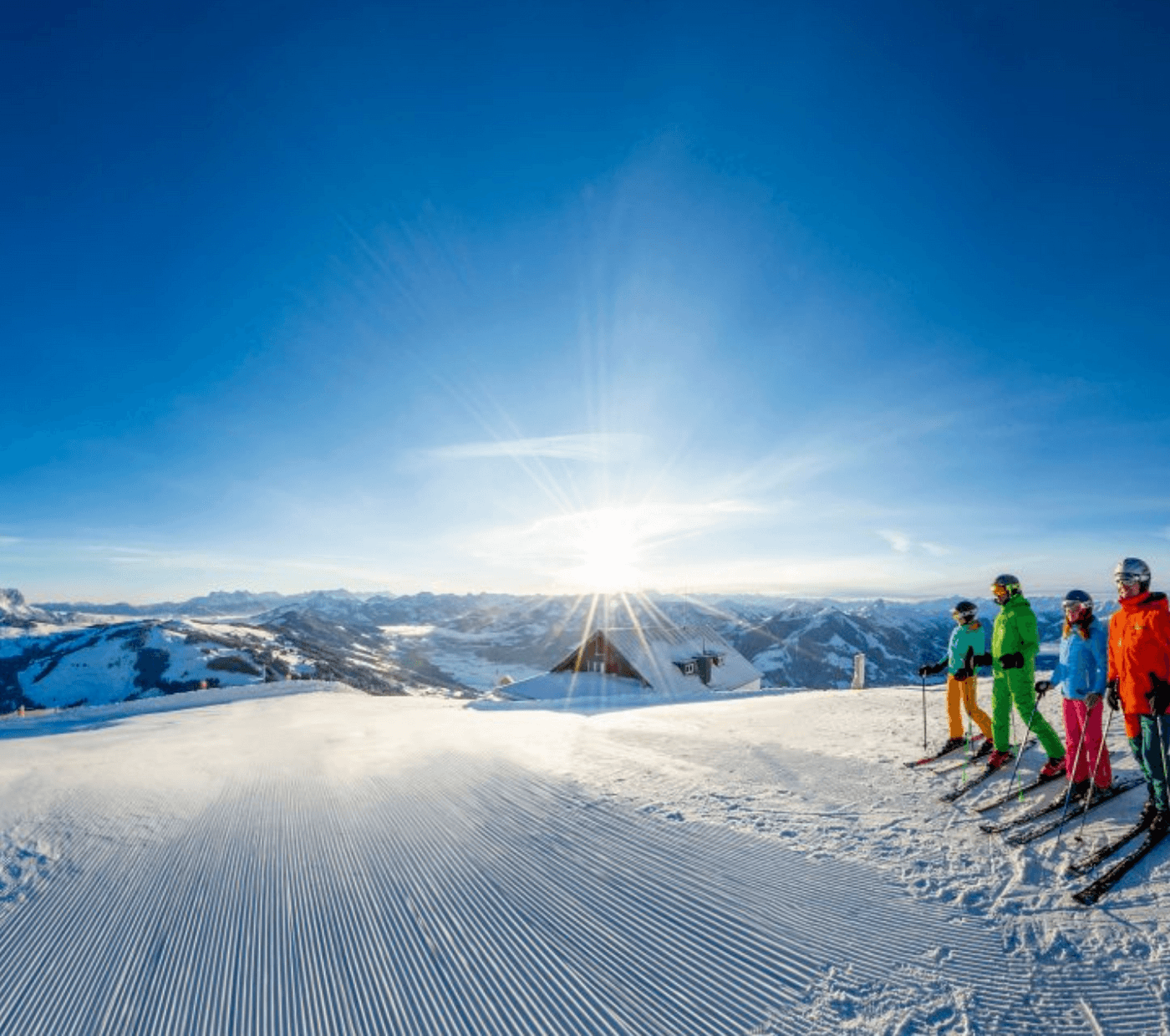 Fyra skidåkare i färgglada kläder står på en preparerad snöig backe med utsikt över bergstoppar och en ljus soluppgång; en liten byggnad syns till höger och fångar magin hos skidresor i Westendorf.