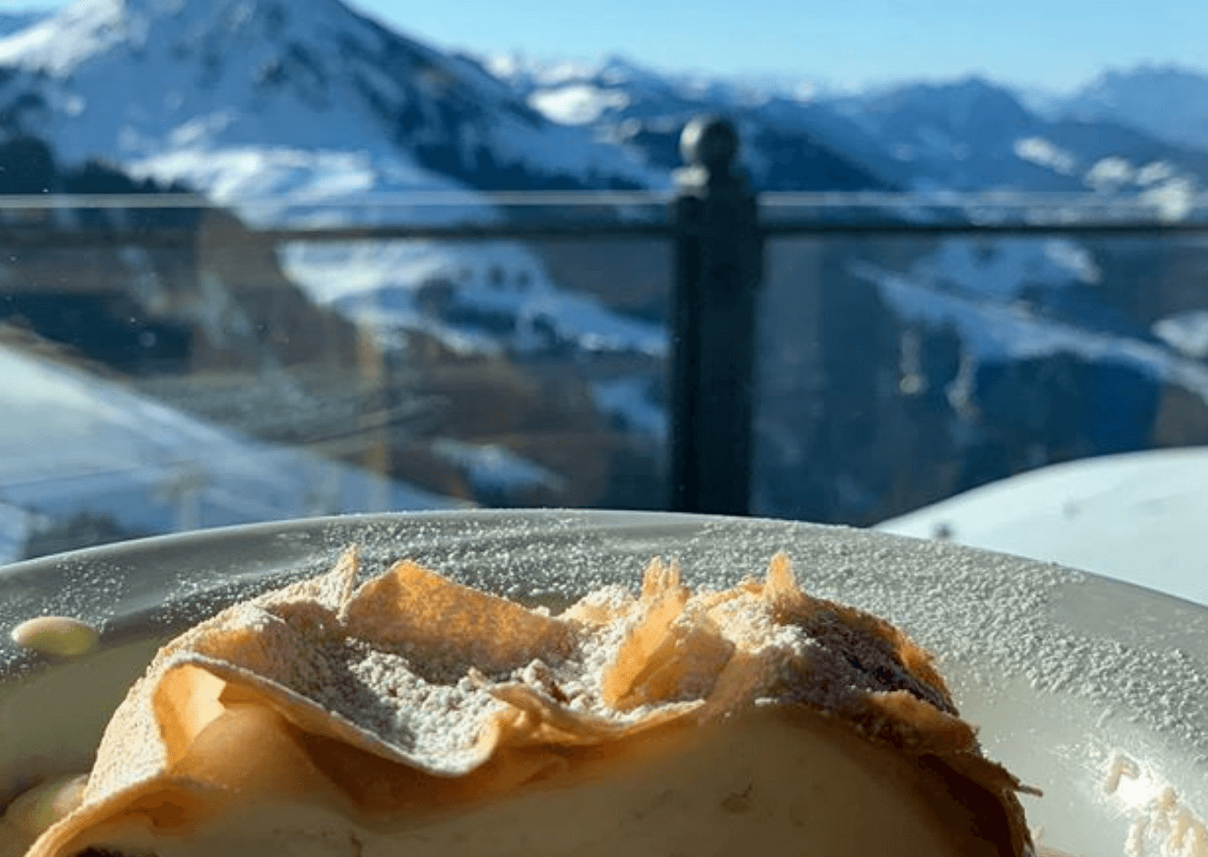 En skive dessert, eventuelt et wienerbrød eller kage, drysset med flormelis på en tallerken. I baggrunden er de snedækkede bjerge i Saalbach synlige under en klar blå himmel, så det føles som et perfekt skiferie øjeblik.