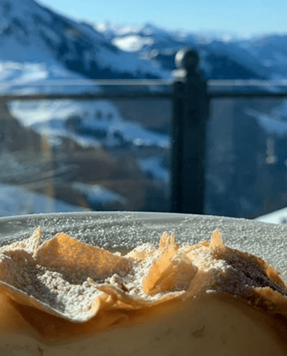 En skive dessert, eventuelt et wienerbrød eller kage, drysset med flormelis på en tallerken. I baggrunden er de snedækkede bjerge i Saalbach synlige under en klar blå himmel, så det føles som et perfekt skiferie øjeblik.