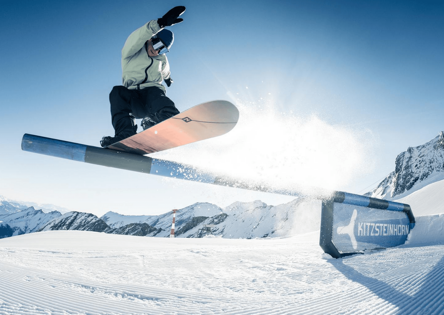 En snowboarder i en let jakke udfører et hop over en skinne med "Kitzsteinhorn" skrevet på den, sat mod den klare himmel og sneklædte bjergterræn. Sne sprøjter bag brættet og fanger spændingen ved skirejser i dette fantastiske alpine landskab.