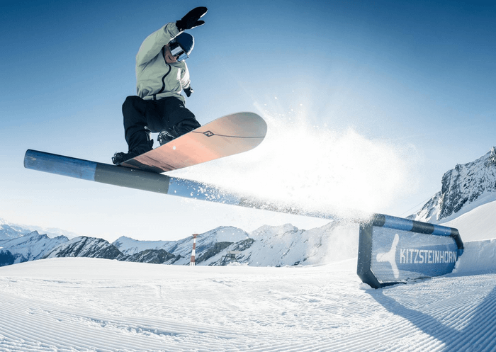 En snowboarder i en let jakke udfører et hop over en skinne med "Kitzsteinhorn" skrevet på den, sat mod den klare himmel og sneklædte bjergterræn. Sne sprøjter bag brættet og fanger spændingen ved skirejser i dette fantastiske alpine landskab.