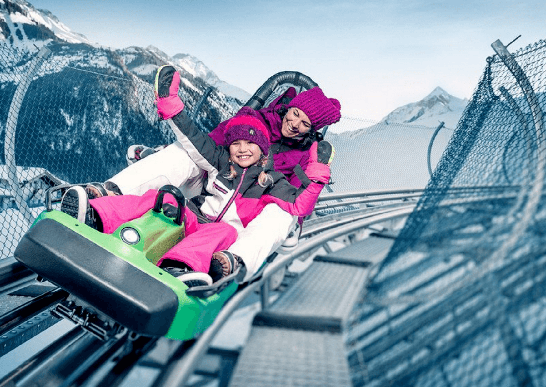 To mennesker bundtet i vintertøj kører på en grøn alpine coaster, smilende og rækker hænderne op. Sneklædte bjerge og et sikkerhedsnet hegn er synlige mod den klare blå himmel i Westendorf, og fanger glæden hos skirejser selv off-piste.