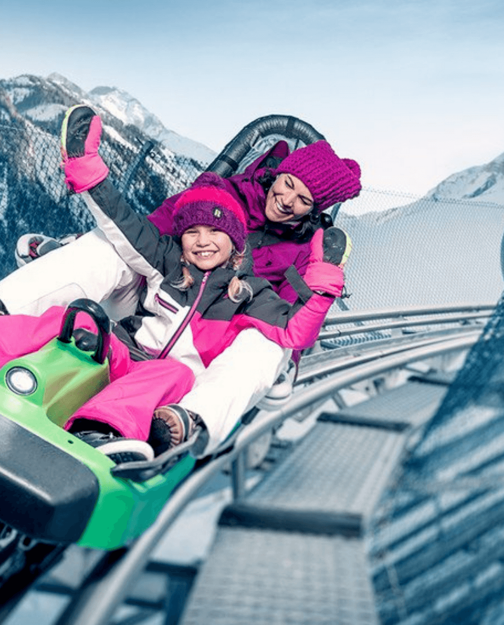 To mennesker bundtet i vintertøj kører på en grøn alpine coaster, smilende og rækker hænderne op. Sneklædte bjerge og et sikkerhedsnet hegn er synlige mod den klare blå himmel i Westendorf, og fanger glæden hos skirejser selv off-piste.