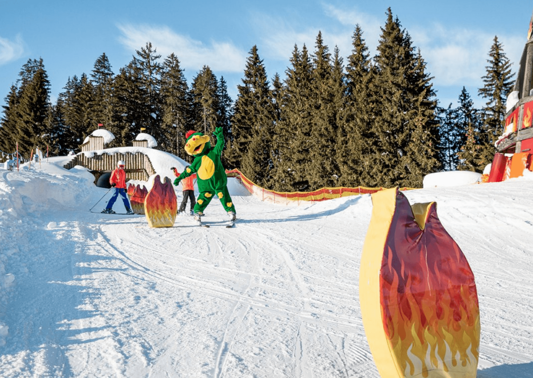 En person i en grön drakekostym åker skidor i en snöig backe under en skidåkning, omgiven av flamformade dekorationer, med andra skidåkare och tallar i bakgrunden under en klarblå himmel.