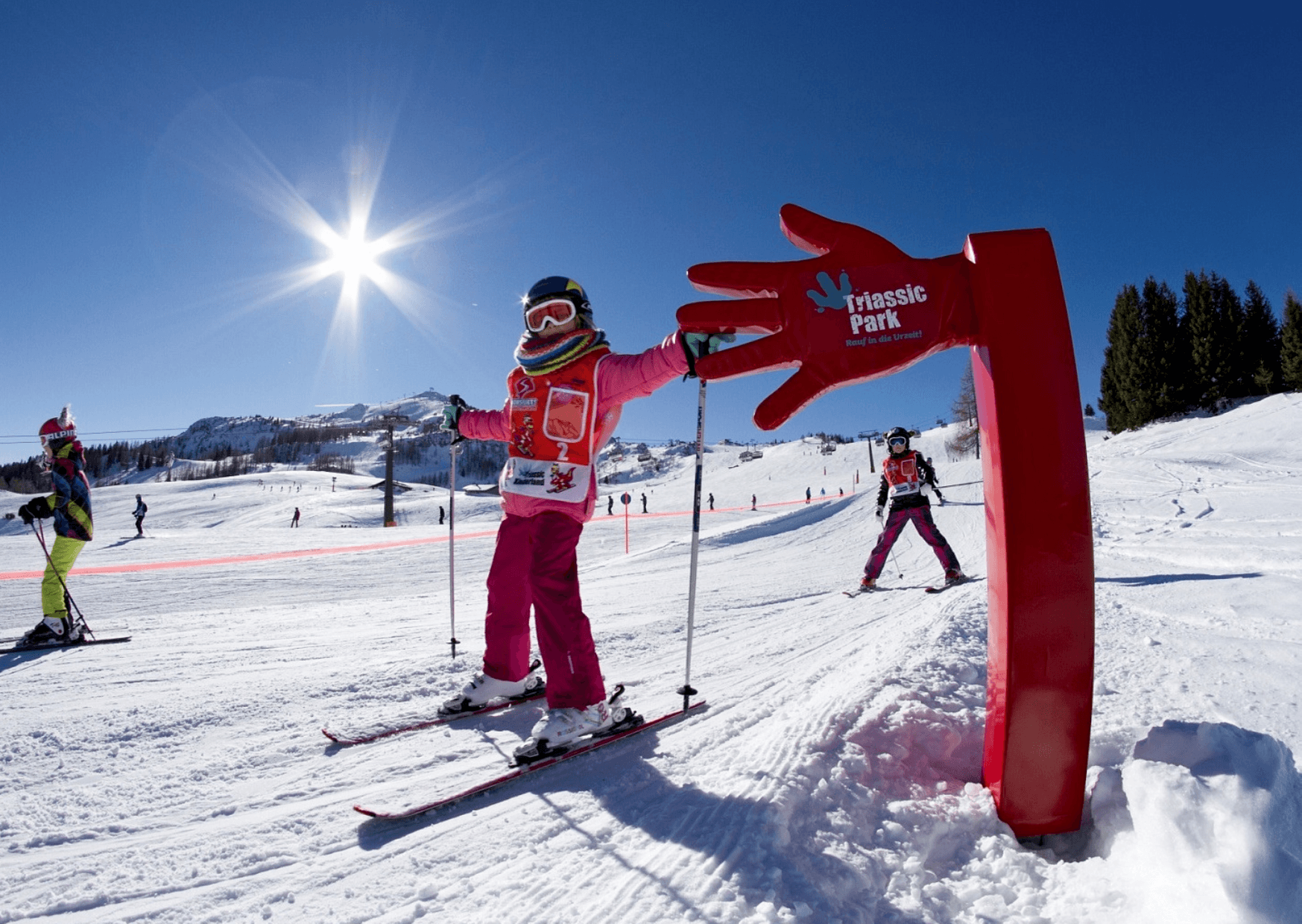 Børn på ski på en solrig, sneklædt skråning med klarblå himmel i Westendorf. Et stort rødt håndformet skilt, hvor der står Triassic Park, skiller sig ud. Scenen er livlig med flere skiløbere og sneklædte bjerge i baggrunden, der fanger charmen ved skirejser i Østrigs vintereventyrland.