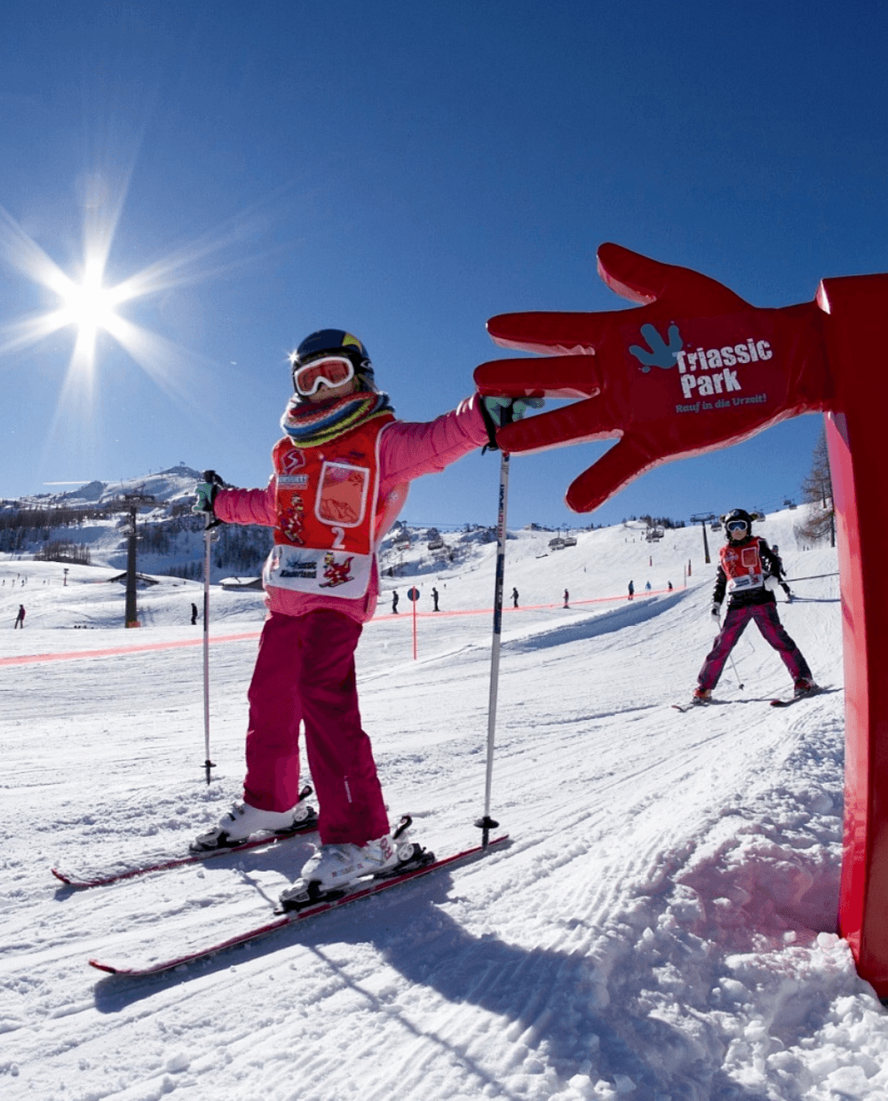 Børn på ski på en solrig, sneklædt skråning med klarblå himmel i Westendorf. Et stort rødt håndformet skilt, hvor der står Triassic Park, skiller sig ud. Scenen er livlig med flere skiløbere og sneklædte bjerge i baggrunden, der fanger charmen ved skirejser i Østrigs vintereventyrland.