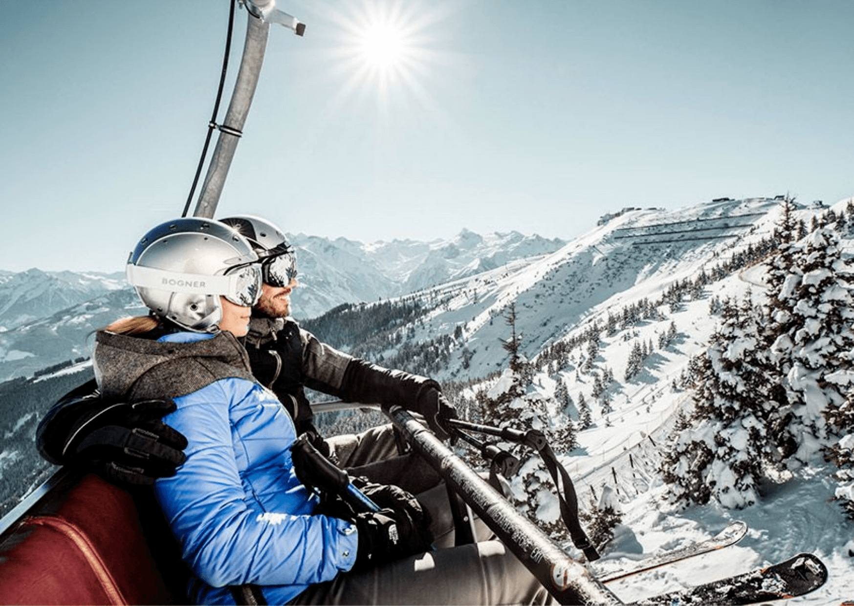 Två personer i vinterkläder och hjälmar åker skidlift uppför ett snötäckt berg i Westendorf, omgivna av snötäckta träd och avlägsna toppar – en perfekt scen för oförglömliga skidresor en klar dag.
