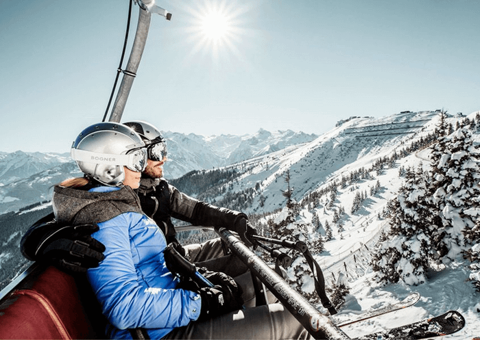 Två personer i vinterkläder och hjälmar åker skidlift uppför ett snötäckt berg i Westendorf, omgivna av snötäckta träd och avlägsna toppar – en perfekt scen för oförglömliga skidresor en klar dag.