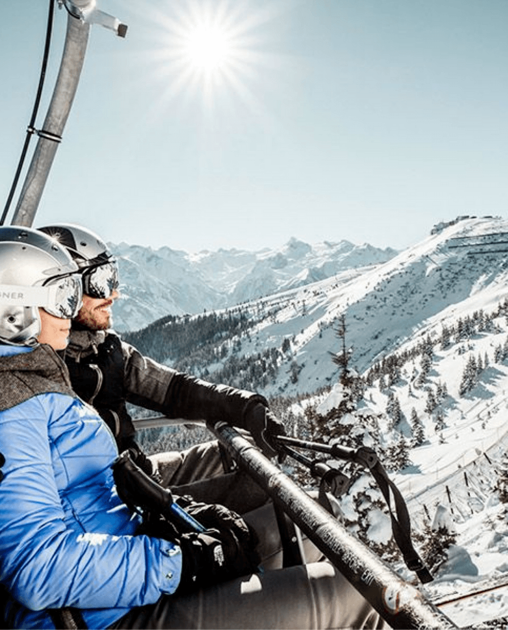 Två personer i vinterkläder och hjälmar åker skidlift uppför ett snötäckt berg i Westendorf, omgivna av snötäckta träd och avlägsna toppar – en perfekt scen för oförglömliga skidresor en klar dag.