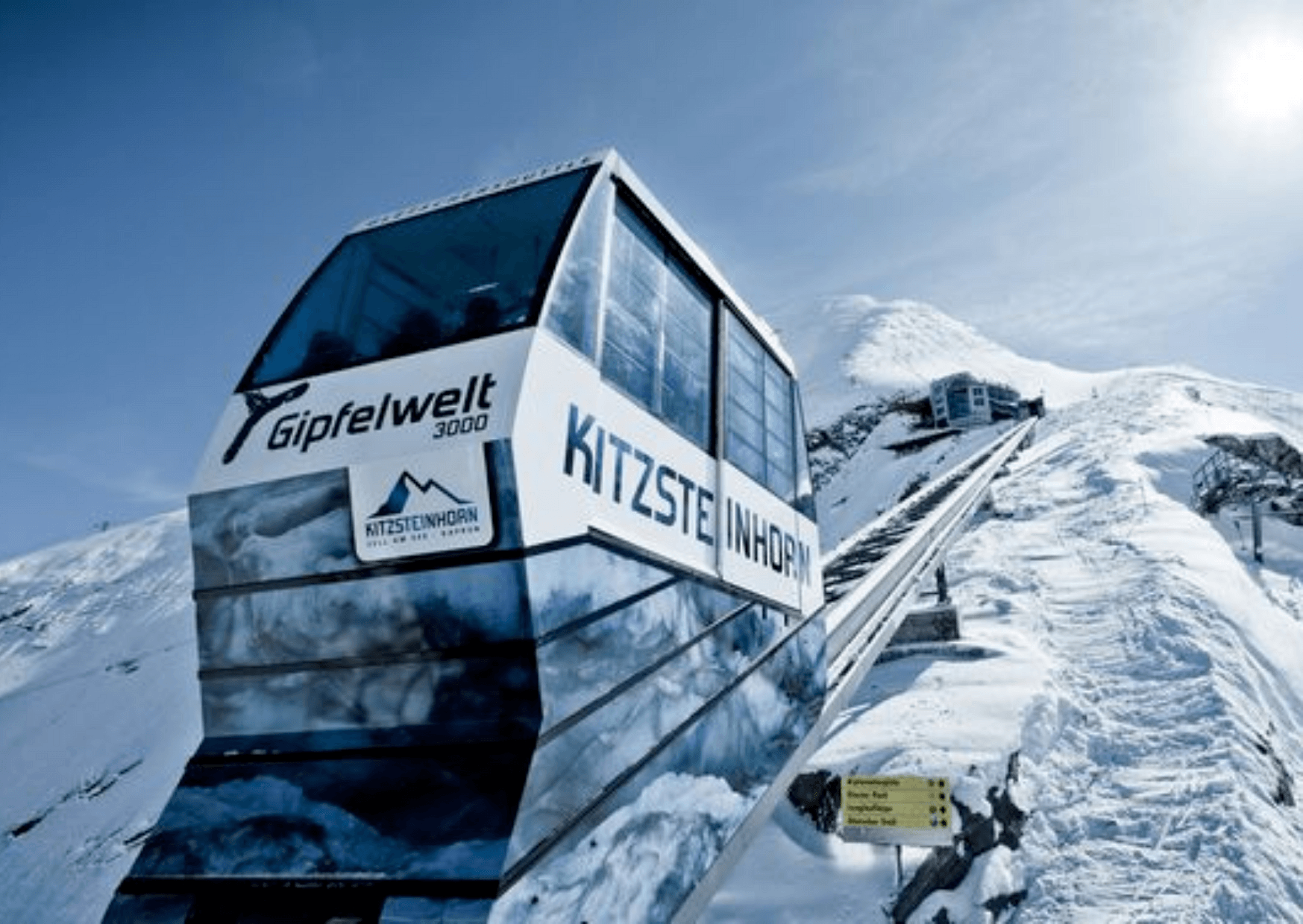 En svævebane mærket Kitzsteinhorn Gipfelwelt 3000 bestiger et snedækket bjerg under en klar blå himmel, der minder om de maleriske landskaber, man kan finde på skirejsedestinationer som Westendorf. Snedækkede tinder og en bjergstation pryder baggrunden med et gult retningsskilt ved siden af banen.