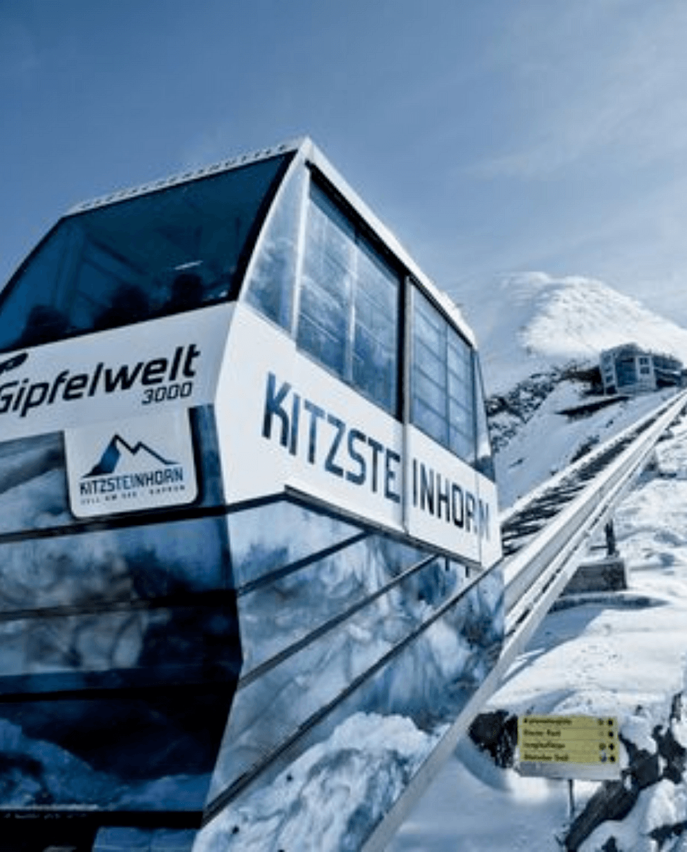 En svævebane mærket Kitzsteinhorn Gipfelwelt 3000 bestiger et snedækket bjerg under en klar blå himmel, der minder om de maleriske landskaber, man kan finde på skirejsedestinationer som Westendorf. Snedækkede tinder og en bjergstation pryder baggrunden med et gult retningsskilt ved siden af banen.