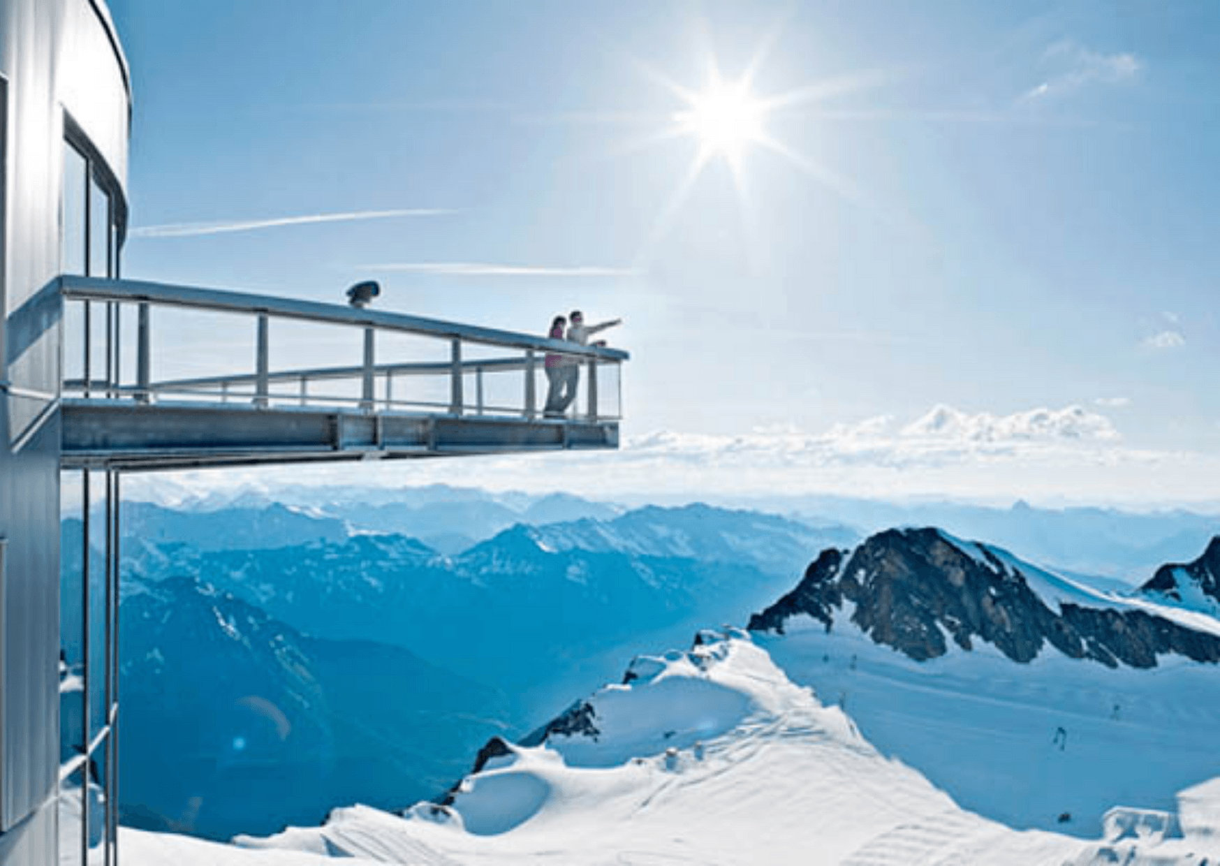 To personer står på en udsigtsplatform, der strækker sig fra en snedækket bygning i Saalbach, med udsigt over en panoramisk bjergkæde under en klar blå himmel og sol. Snedækkede tinder og en stor himmel skaber den fredfyldte, betagende udsigt, der er typisk for skirejseoplevelser.