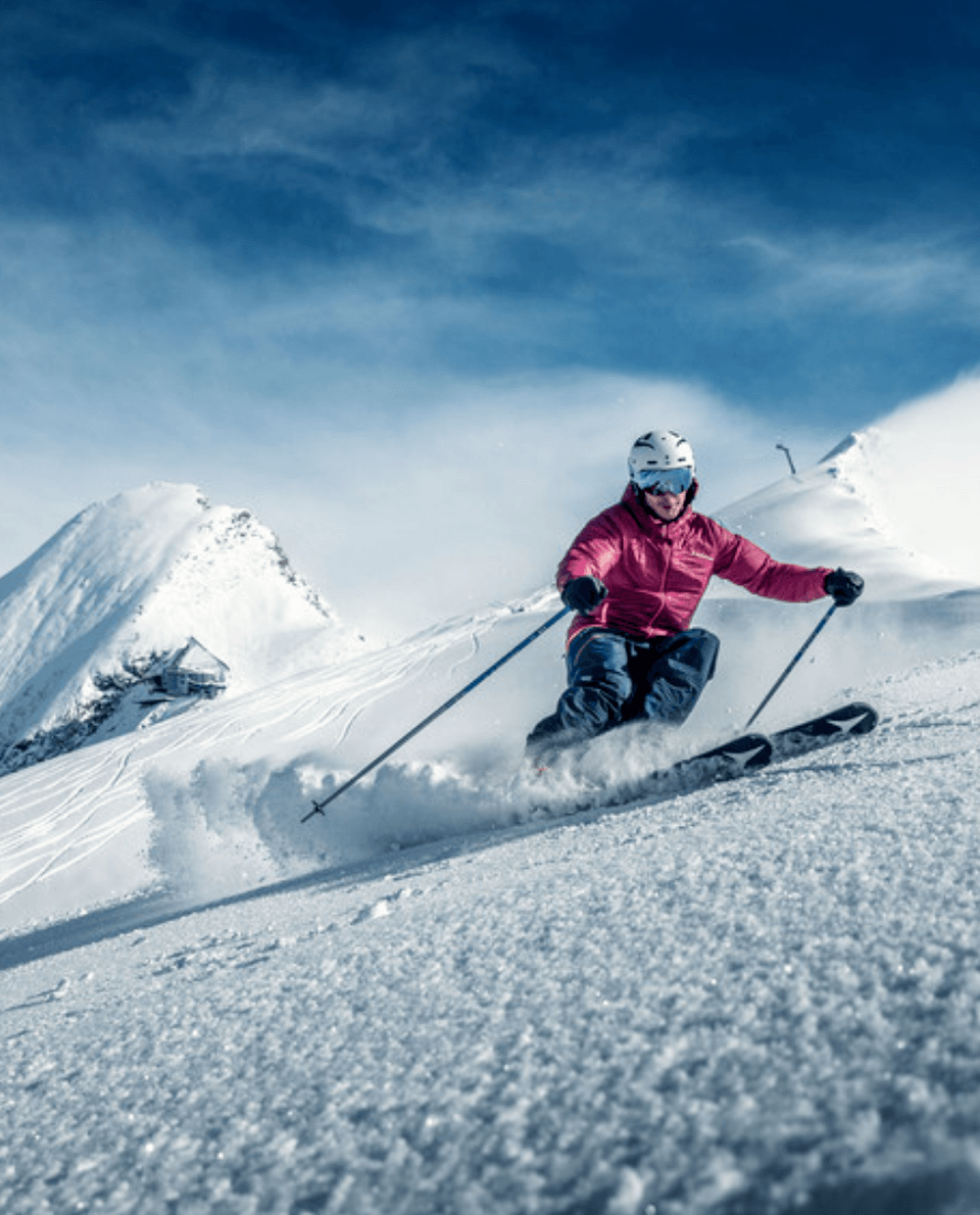 En skidåkare i röd jacka och hjälm åker snabbt nerför en snöig Saalbach-bergssluttning och lämnar ett spår av puder, med snötäckta toppar och en blå himmel i bakgrunden.