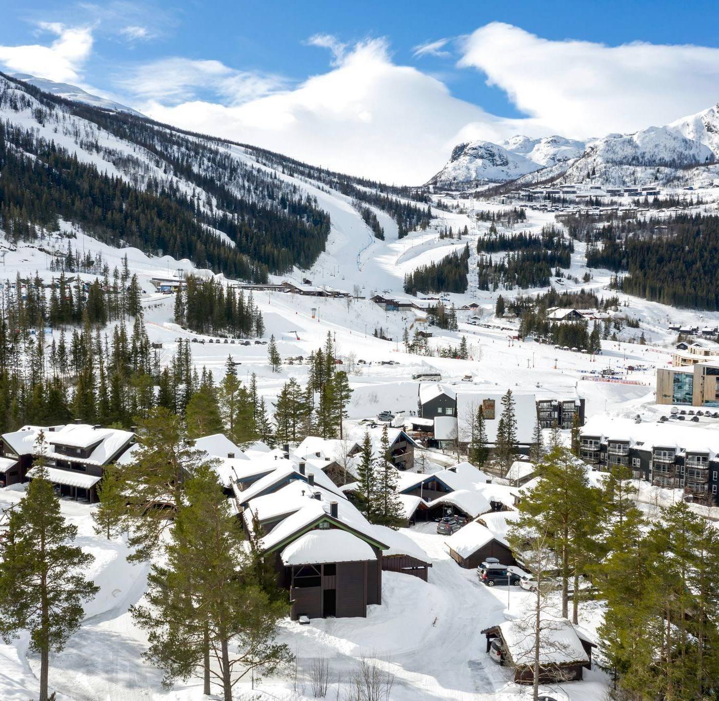 Et sneklædt bjerglandskab i Westendorf har et skisportssted med hytter beliggende blandt stedsegrønne træer. Skiløjper og stier er synlige i baggrunden under en klar blå himmel med skyer, perfekt til en uforglemmelig skiferie.
