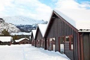 En række mørke træhytter med røde vinduer er dækket af sne og ligger i et snedækket landskab med bjerge i baggrunden under en delvis overskyet himmel.