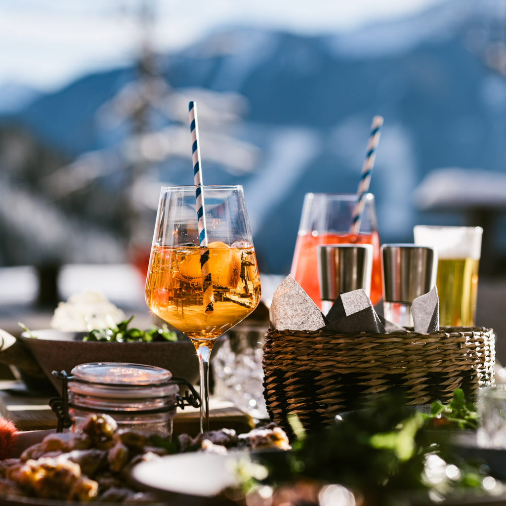 Et bord dækket til udendørs nydelse med to cocktails garneret med stribede sugerør, en kurv med metalkopper og snacks