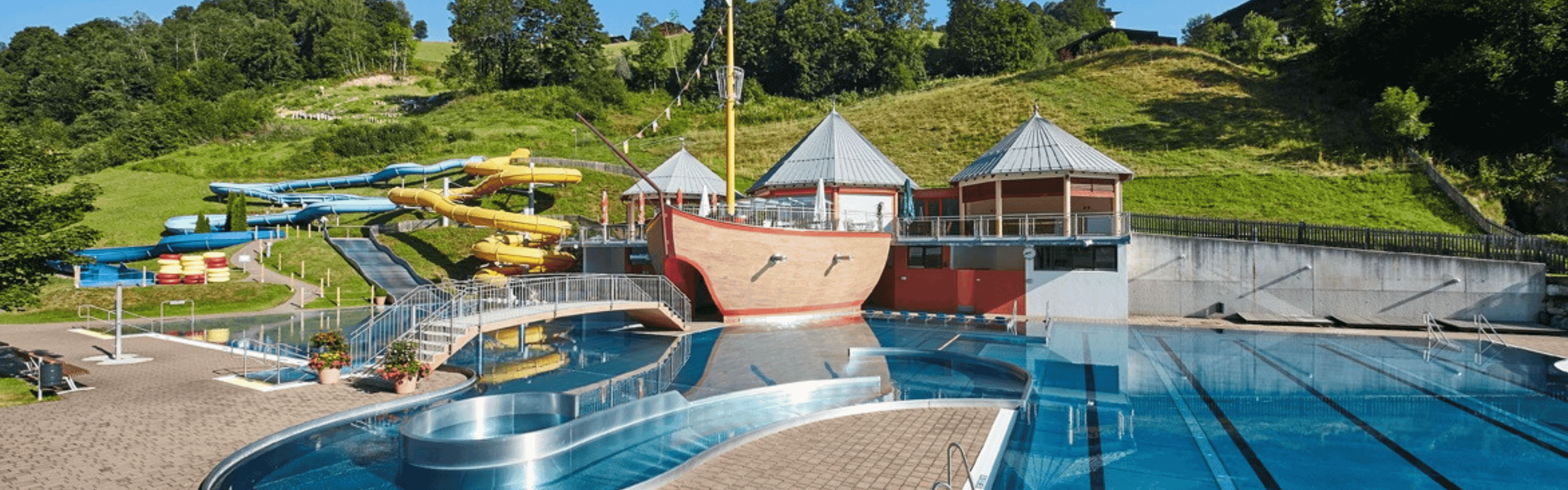 Udendørs vandland med en stor pool med blåt vand, en bro og en central struktur med piratskibstema. Forskellige farverige vandrutsjebaner snor sig ned ad en græsklædt bjergskråning i baggrunden. Træer og en klar himmel er synlige.