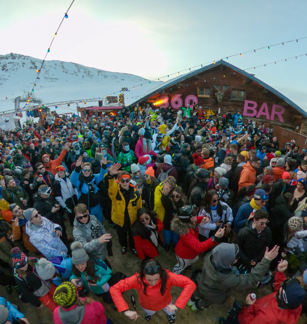 Et livligt publikum på en udendørs bar i sneklædte bjergomgivelser, der fejrer Nytår på ski. Folk er klædt i farverigt