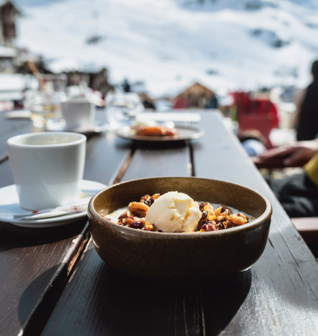 En skål dessert med is og nødder står på et træbord udendørs, omgivet af snedækkede bjerge, og danner den perfekte