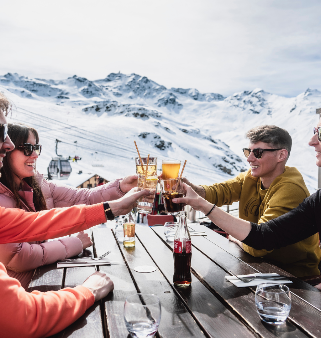 Fire personer skåler med drinks ved et udendørs træbord mod en sneklædt bjergkulisse, der fejrer nytår på ski. Klædt i