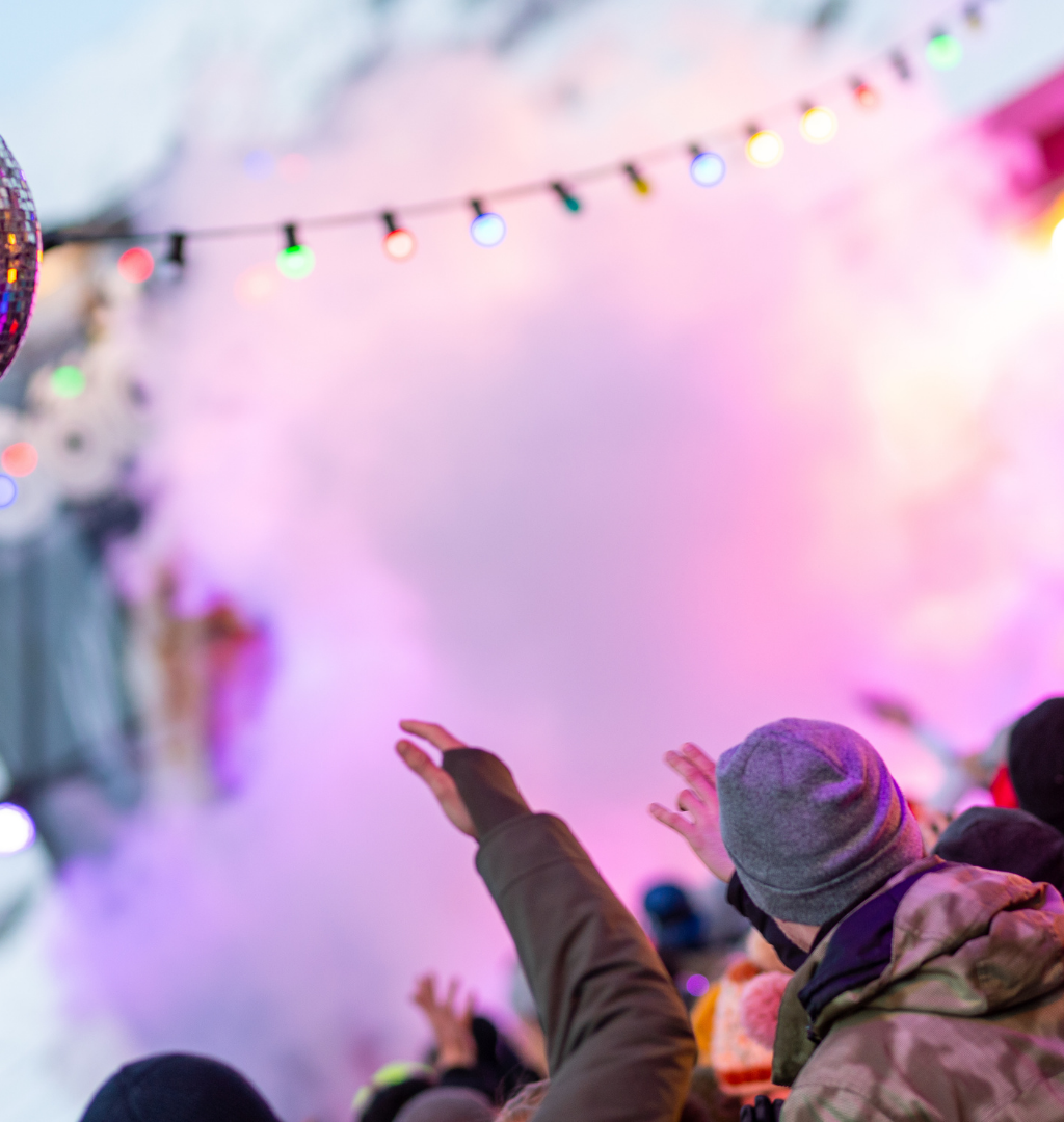 Et livligt publikum klædt i vintertøj nyder en udendørs fest, der minder om Nytår på ski. Klare, farverige lys glimter