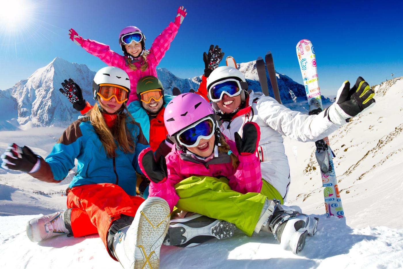 En grupp på fem personer i färgglada skidkläder sitter på en snöig backe i Saalbach och ler och höjer armarna. Snötäckta berg och en klarblå himmel fungerar som bakgrund. Skidor och skidstavar står uppsatta i närheten och fångar det perfekta skidreseögonblicket.