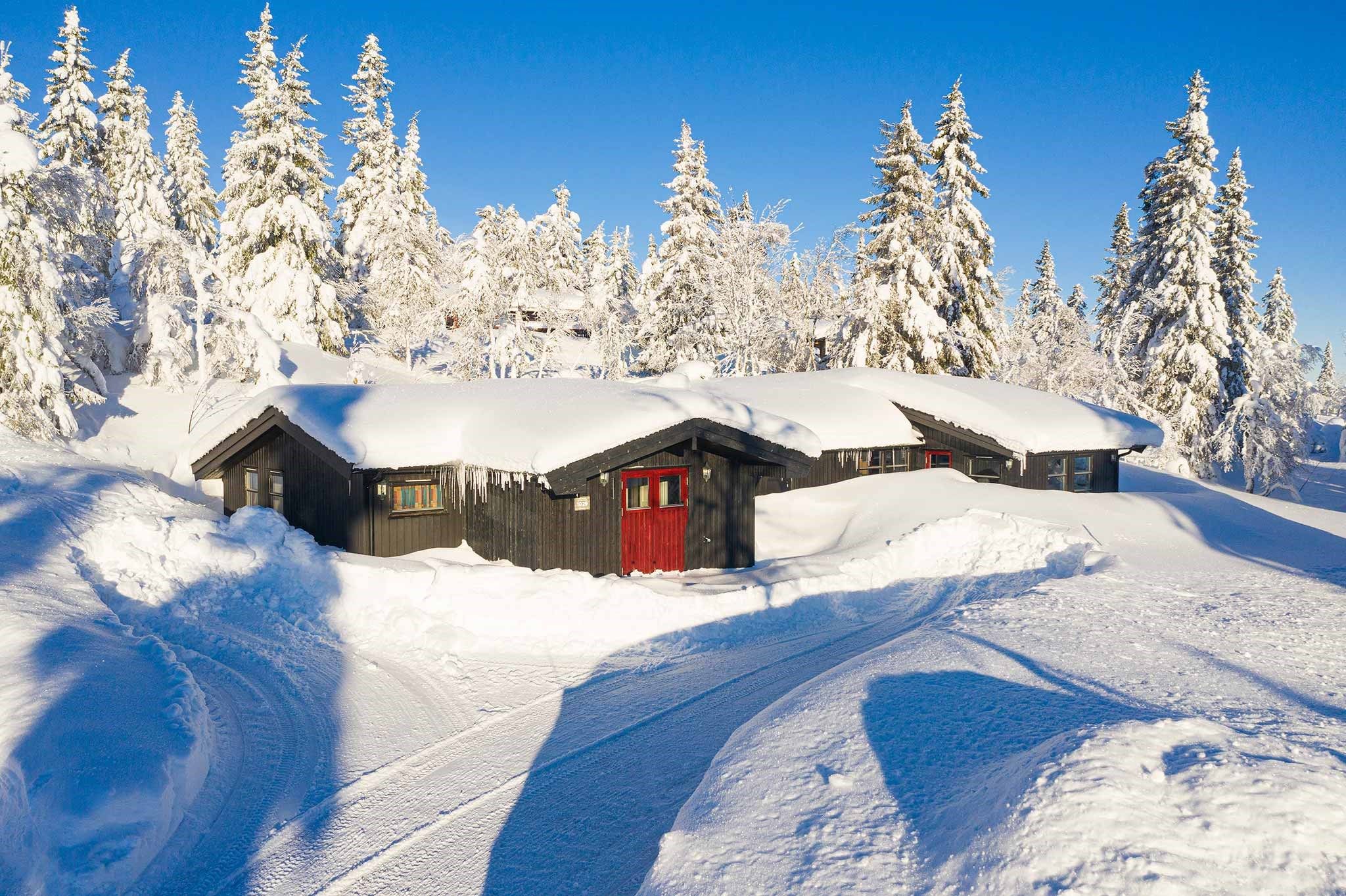 En hyggelig hytte med røde døre ligger i det snedækkede landskab i Westendorf, omgivet af snefyldte træer under en klar blå himmel. Dækspor er synlige i sneen, hvilket fører til denne perfekte skiferieferie.
